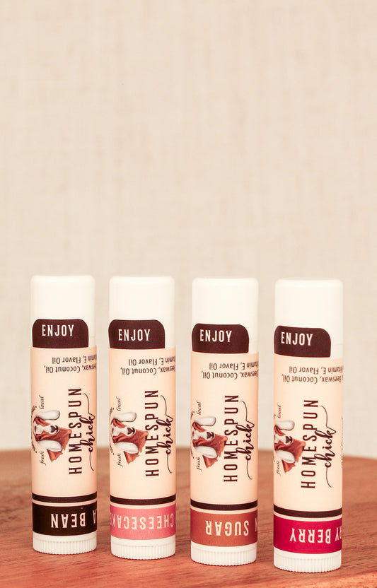 Four lip balm tubes standing upright on a wooden surface against a light beige textured background. The tubes are labeled ENJOY HOMESPUN chick in flavors Vanilla Bean, Strawberry Cheesecake, Brown Sugar, and Cherry Berry, with cream-colored bodies, dark brown labels, and pink, brown, and red accents.